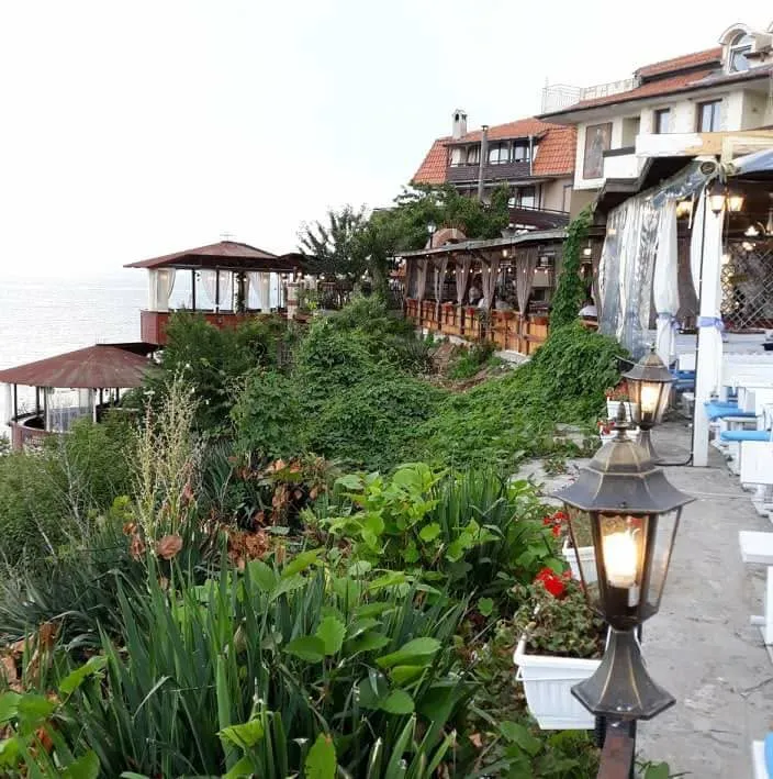 The clifftop setting of Restaurant Breeze in Nessebar Old Town: tiered wooden terraces and gazebos above the water, dense greenery, an iron lantern, and the Black Sea stretching away to the left.