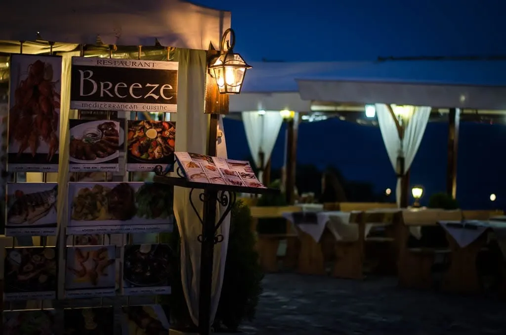 Restaurant Breeze at dusk: the lit sign reading Restaurant Breeze, Mediterranean cuisine, beside a lantern and an illustrated menu board, with white-draped tables set on the terrace behind and the sea going dark.