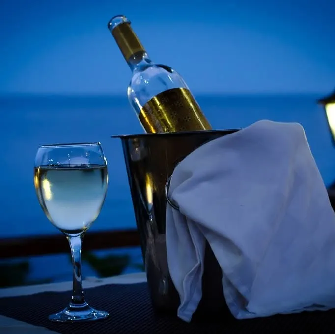 A bottle of white wine in a steel ice bucket with a white napkin, and a filled glass beside it, on a terrace table at Restaurant Breeze with the Black Sea deep blue behind at dusk.